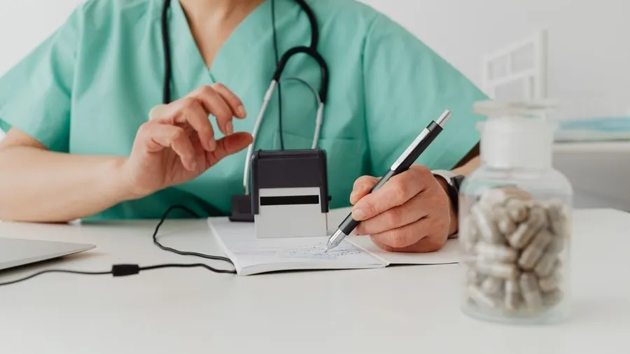 Practitioner in scrubs writing notes at a clinic desk beside a jar of supplies