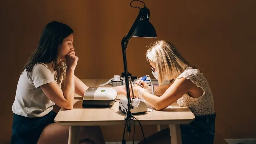 Two people at a nail desk with a lamp in a warm-toned salon