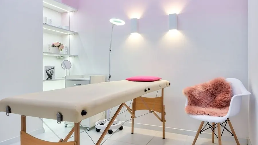 Bright facial treatment room with a padded bed, lamp and shelving