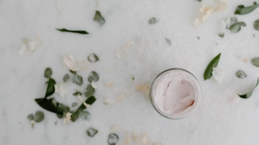 Open jar of pink cream on a marble surface scattered with dried petals and leaves