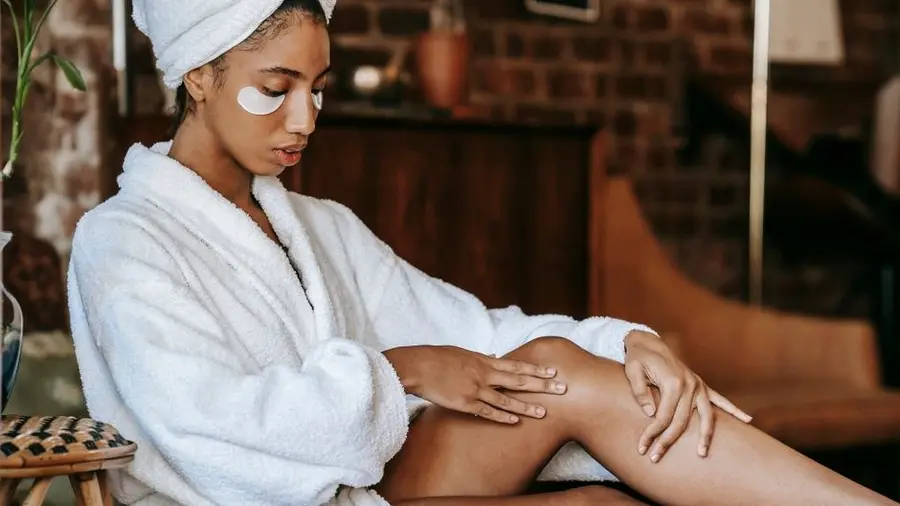 Woman in a bathrobe applying lotion to her legs at home
